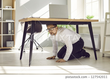 Portrait of a scared stressed young business man hiding under the desk on his workplace at office. Portrait of a scared stressed young business man hiding under the desk on his workplace at office. 127542011