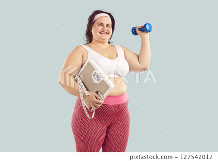 Happy fat woman wearing sportswear holding scales, dumbbell and measuring tape on blue background. Happy fat woman wearing sportswear holding scales, dumbbell and measuring tape on blue background. 127542012