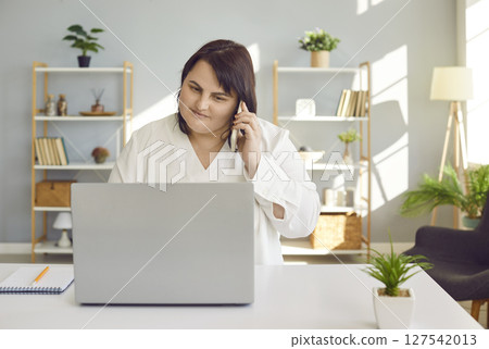 Plus size woman sitting at laptop and having phone conversation in office. Plus size woman sitting at laptop and having phone conversation in office. 127542013
