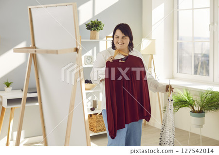 Happy overweight young woman choosing between two blouses, looking in mirror and smiling Happy overweight young woman choosing between two blouses, looking in mirror and smiling 127542014