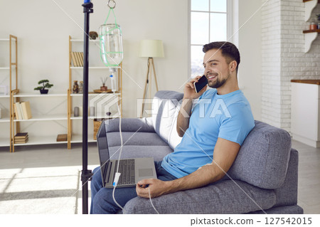Young man talking by mobile phone sitting on sofa at home while receiving IV vitamin therapy. Young man talking by mobile phone sitting on sofa at home while receiving IV vitamin therapy. 127542015