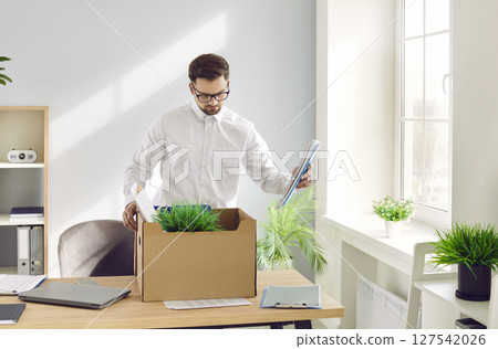 Sad, fired young corporate employee packing stuff off his desk in a cardboard box Sad, fired young corporate employee packing stuff off his desk in a cardboard box 127542026