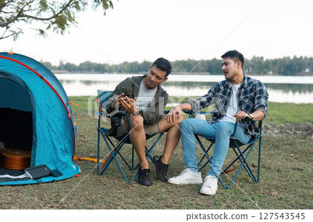 Gay LGBTQIA couple sitting on picnic chair drinking tea and coffee while camping on vacation holiday Gay LGBTQIA couple sitting on picnic chair drinking tea and coffee while camping on vacation holiday 127543545
