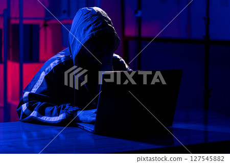 A man in a black hoodie intently using a laptop in a neon-lit room 127545882