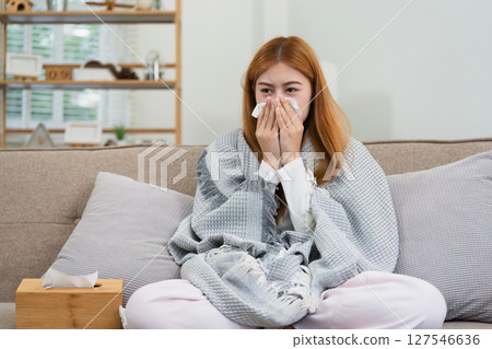 young woman sitting on sofa covered with blanket freezing blowing running nose got fever caught cold sneezing in tissue young woman sitting on sofa covered with blanket freezing blowing running nose got fever caught cold sneezing in tissue 127546636