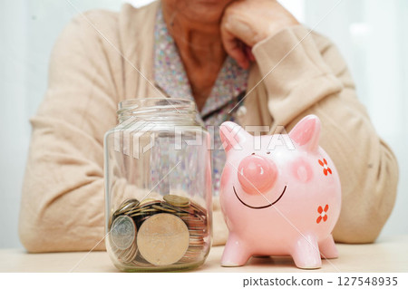 Retired elderly woman counting coins money with piggy bank and worry about monthly expenses and treatment fee payment. 127548935