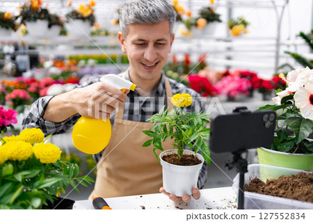 A man in a flower shop waters a plant while filming a tutorial with his phone, teaching online. 127552834