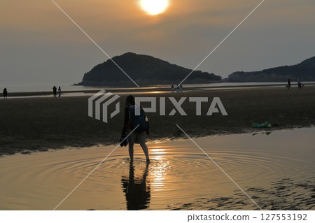 A woman wading through the tide pools at Chichibugahama Beach in Mitoyo, with the setting sun reflecting off the beach 127553192