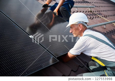 Workers building solar panel system on rooftop of house. Two men installers in helmets installing photovoltaic solar module outdoors. Alternative, green and renewable energy generation concept. 127553998