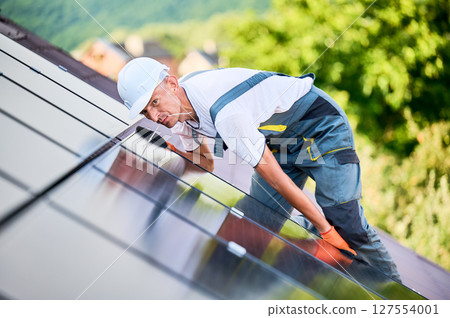 Worker building photovoltaic solar panel system on roof of house. Engineer in helmet looking on evenness of solar modules during installing outdoors. Concept of alternative and renewable energy. 127554001