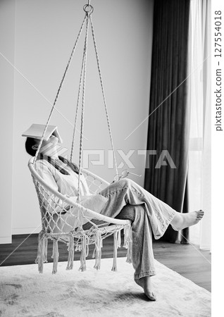 Tired girl fall asleep after reading a book. Young female relaxing in hanging chair after hard day. Pretty girl lying in swing with book on her face. Black and white image. 127554018