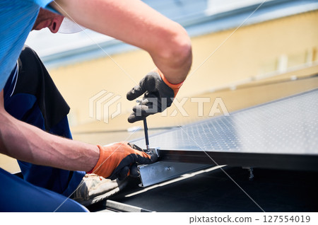 Worker building photovoltaic solar panel system on rooftop of house. Close up of man engineer in gloves installing solar module with help of hex key outdoors. Renewable energy generation concept. 127554019