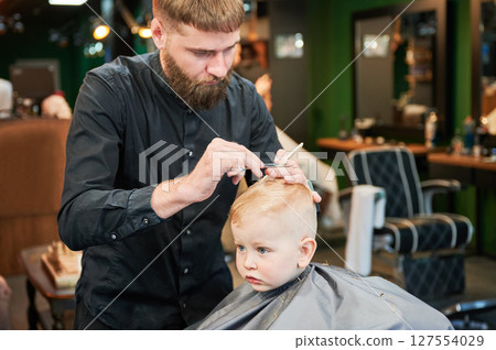 Man barber cutting little boy's hair using comb and scissors. Child getting haircut from adult male, likely barber. Professional hairdresser and cute client at modern barbershop. 127554029