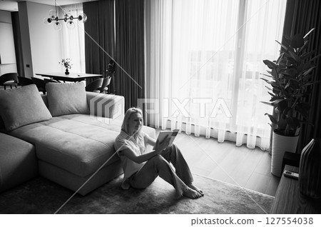 Young woman sitting next to couch and reading. Beautiful female relaxing on wooden floor. Woman sitting with book opposite window in natural light. Black and white image. Young woman sitting next to couch and reading. Beautiful female relaxing on wooden floor. Woman sitting with book opposite window in natural light. Black and white image. 127554038