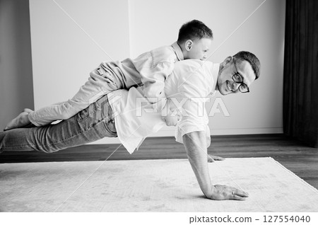 Energetic father and his little son training at home. Kid climbed dad's back while he doing plank. Young family enjoying activity games at home. Black and white image. 127554040