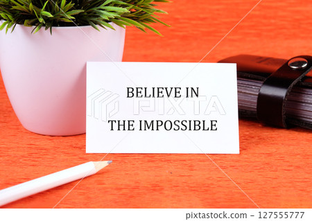 Motivational quote. BELIEVE IN THE IMPOSSIBLE text written on a card next to a small potted plant and stationery Motivational quote. BELIEVE IN THE IMPOSSIBLE text written on a card next to a small potted plant and stationery 127555777