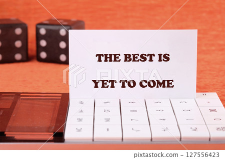 Motivational quotes THE BEST IS YET TO COME on a business card with game cubes in the background and a calculator in the foreground Motivational quotes THE BEST IS YET TO COME on a business card with game cubes in the background and a calculator in the foreground 127556423