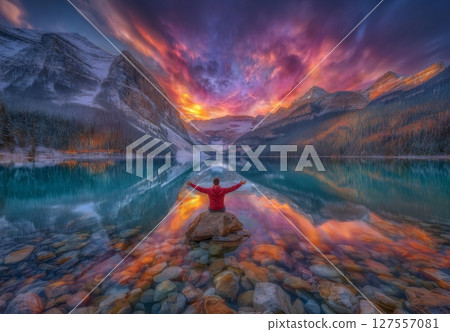 Seated on a rock, a man in a red outfit gazes out at the morning clouds and their reflections on the surface of Lake Louise 127557081