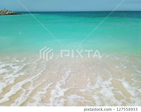 Beautiful view of the UAE s pristine coastline, where gentle sea waves lap against soft, powdery white sand. The crystal-clear turquoise waters create a serene contrast with the bright, sunlit beach. 127558933