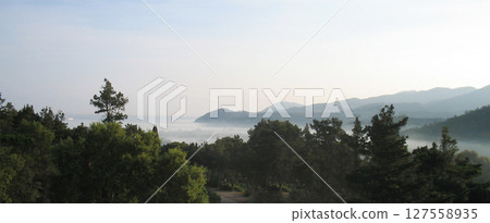 The soft glow of morning sun rays gently breaks through a light haze, casting a warm, ethereal light over the rolling hills and towering mountains of Elba Island in spring. The delicate mist drapes. The soft glow of morning sun rays gently breaks through a light haze, casting a warm, ethereal light over the rolling hills and towering mountains of Elba Island in spring. The delicate mist drapes. 127558935