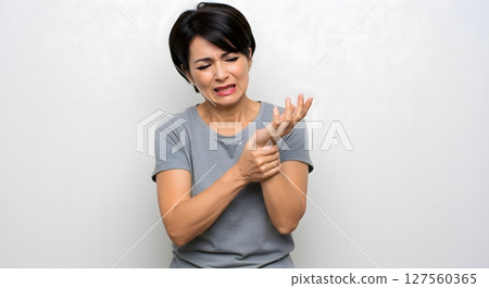 Asian middle aged woman in gray shirt holding wrist in pain with grimace indoors Asian middle aged woman in gray shirt holding wrist in pain with grimace indoors 127560365
