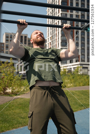 Sporty blonde caucasian bearded man does pull ups on horizontal bar at urban sports ground. Summer. Workout. Morning routine. 127561934
