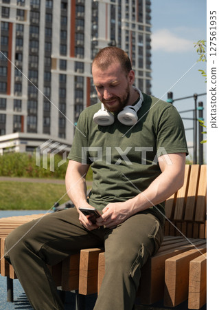 Portrait of caucasian bearded man with white headphones relax and sitting on bench with smartphone on urban sports ground. Outdoors. Summer season. 127561935