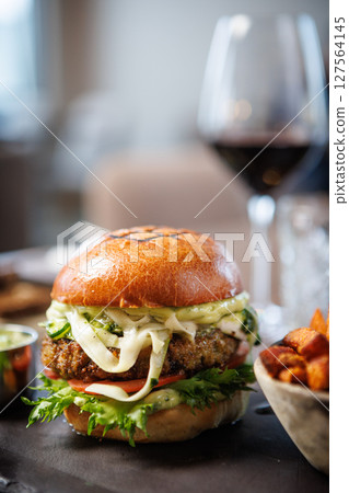 Gourmet veggie burger with lettuce, tomato, cheese, and sauce on a brioche bun. Served with a side of crispy sweet potato fries and a small cup of sauce. Vegetarian meal served in modern restaurant 127564145