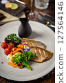 Pan-seared fish fillet with vibrant vegetables, including broccoli, cherry tomatoes, and carrot ribbons, garnished with edible flowers. Accompanied by a dark sauce jug. Fine dining closeup 127564146