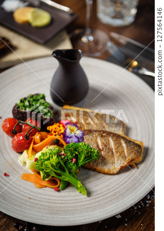 Pan-seared fish fillet with vibrant vegetables, including broccoli, cherry tomatoes, and carrot ribbons, garnished with edible flowers. Accompanied by a dark sauce jug. Fine dining closeup 127564146