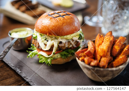 Gourmet veggie burger with lettuce, tomato, cheese, and sauce on a brioche bun. Served with a side of crispy sweet potato fries and a small cup of sauce. Vegetarian meal served in modern restaurant Gourmet veggie burger with lettuce, tomato, cheese, and sauce on a brioche bun. Served with a side of crispy sweet potato fries and a small cup of sauce. Vegetarian meal served in modern restaurant 127564147
