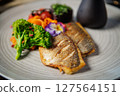 Crispy fish fillet paired with fresh broccoli, cherry tomatoes, and carrot ribbons, topped with colorful edible flowers. Served with a dark sauce jug. Elegant and healthy meal presentation 127564151