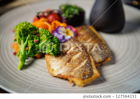 Crispy fish fillet paired with fresh broccoli, cherry tomatoes, and carrot ribbons, topped with colorful edible flowers. Served with a dark sauce jug. Elegant and healthy meal presentation 127564151