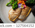 Pan-seared fish fillet with vibrant vegetables, including broccoli, cherry tomatoes, and carrot ribbons, garnished with edible flowers. Accompanied by a dark sauce jug. Fine dining closeup 127564152