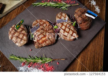 Juicy grilled steaks with Brazilian, Uruguayan, and Estonian flags. Served with fresh herbs and microgreens on a slate plate, showcasing international gourmet cuisine in a fine dining setting 127564153