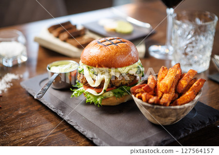 Vegetarian burger with fresh greens, tomato, cheese, and creamy sauce on a brioche bun. Accompanied by a bowl of sweet potato fries and a metal cup of dip. Healthy and tasty dining option 127564157