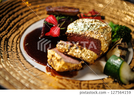 Luxurious gold-plated beef steak accompanied by grilled zucchini, cherry tomatoes, and broccoli. Finished with a rich sauce and edible flowers, presented on a stylish plate in a high-end restaurant Luxurious gold-plated beef steak accompanied by grilled zucchini, cherry tomatoes, and broccoli. Finished with a rich sauce and edible flowers, presented on a stylish plate in a high-end restaurant 127564159
