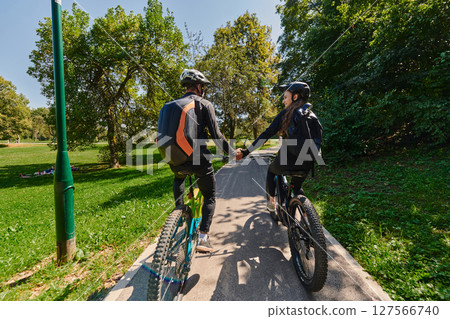 A sweet couple, adorned in cycling gear, rides their bicycles, their hands interlocked in a romantic embrace, capturing the essence of love, adventure, and joy on a sunlit path 127566740