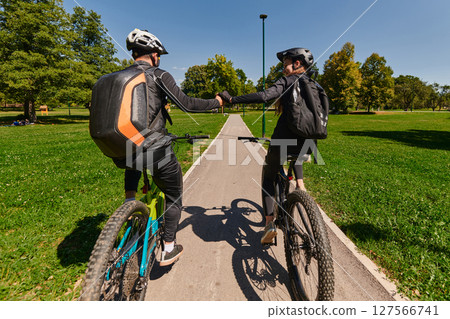 A sweet couple, adorned in cycling gear, rides their bicycles, their hands interlocked in a romantic embrace, capturing the essence of love, adventure, and joy on a sunlit path 127566741