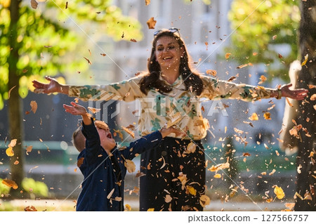 A modern woman joyfully plays with her son in the park, tossing leaves on a beautiful autumn day, capturing the essence of family life and the warmth of mother-son bonding in the midst of the fall 127566757