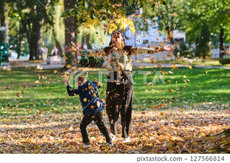 A modern woman joyfully plays with her son in the park, tossing leaves on a beautiful autumn day, capturing the essence of family life and the warmth of mother-son bonding in the midst of the fall 127566814