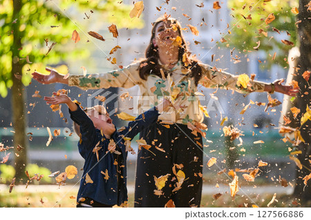 A modern woman joyfully plays with her son in the park, tossing leaves on a beautiful autumn day, capturing the essence of family life and the warmth of mother-son bonding in the midst of the fall 127566886