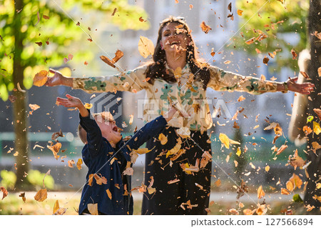 A modern woman joyfully plays with her son in the park, tossing leaves on a beautiful autumn day, capturing the essence of family life and the warmth of mother-son bonding in the midst of the fall 127566894