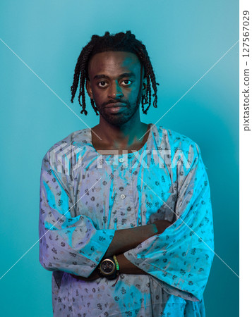 A Sudanese man adorned with modern dreadlocks stands proudly in traditional Sudanese attire, his arms crossed, conveying a blend of cultural heritage and contemporary style against a vibrant blue 127567029