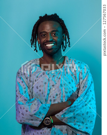 A Sudanese man adorned with modern dreadlocks stands proudly in traditional Sudanese attire, his arms crossed, conveying a blend of cultural heritage and contemporary style against a vibrant blue A Sudanese man adorned with modern dreadlocks stands proudly in traditional Sudanese attire, his arms crossed, conveying a blend of cultural heritage and contemporary style against a vibrant blue 127567033