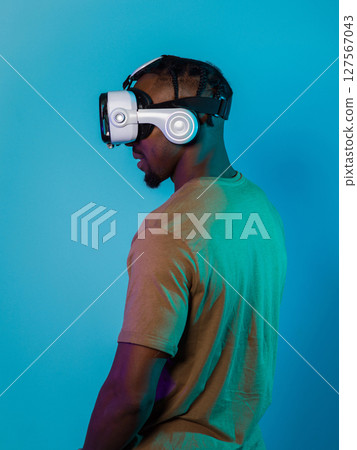 In a futuristic visual, an African American man stands isolated against a striking blue backdrop, adorned with VR glasses that transport him into a cutting-edge virtual reality experience, merging In a futuristic visual, an African American man stands isolated against a striking blue backdrop, adorned with VR glasses that transport him into a cutting-edge virtual reality experience, merging 127567043