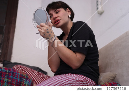 Young woman applying makeup while looking in a handheld mirror at home Young woman applying makeup while looking in a handheld mirror at home 127567216