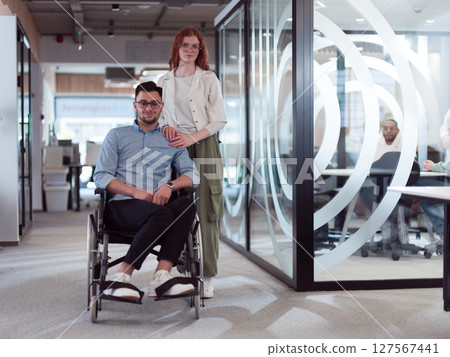 Young business colleagues, collaborative business colleagues, including a person in a wheelchair, walk past a modern glass office corridor, illustrating diversity, teamwork and empowerment in the Young business colleagues, collaborative business colleagues, including a person in a wheelchair, walk past a modern glass office corridor, illustrating diversity, teamwork and empowerment in the 127567441