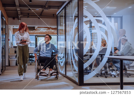 Young business colleagues, collaborative business colleagues, including a person in a wheelchair, walk past a modern glass office corridor, illustrating diversity, teamwork and empowerment in the Young business colleagues, collaborative business colleagues, including a person in a wheelchair, walk past a modern glass office corridor, illustrating diversity, teamwork and empowerment in the 127567853