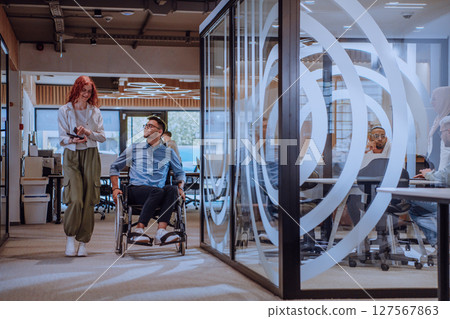 Young business colleagues, collaborative business colleagues, including a person in a wheelchair, walk past a modern glass office corridor, illustrating diversity, teamwork and empowerment in the 127567863